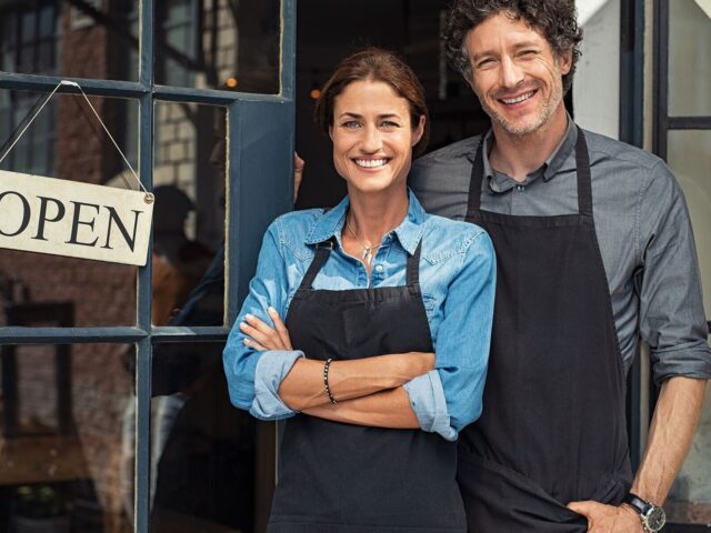 Business owning couple wearing shop aprons stand outside door with 22 Open22 sign 978b8d064138018a1dfad71b9882dd21